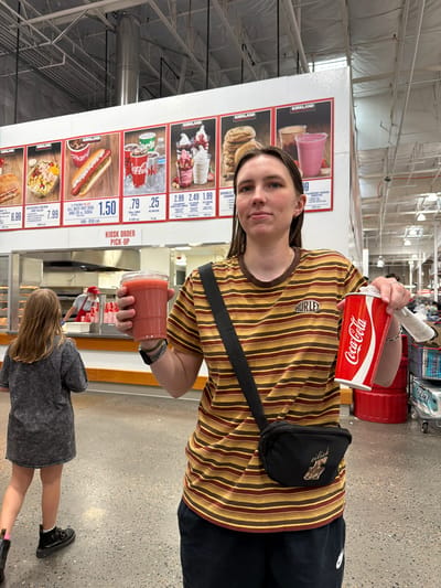 A woman in a striped shirt and fanny pack stands in a Costco food court in Beaverton, Oregon. She holds a red cup of pink smoothie in her left hand and a can of Coca-Cola in her right. In the background, a young girl walks away from the camera, and a Costco employee is visible behind the counter. The food court is brightly lit, with a menu board displaying various food items.