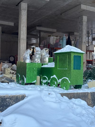 This image, captured in L'vovskiy, Russia, depicts a static scene within an outdoor or semi-enclosed concrete structure, characterized by prominent concrete pillars supporting an overhead concrete slab. The foreground is covered by a substantial, uneven layer of white snow, partially obscuring a dark, textured concrete or stone ledge.
The central subject is a large, two-part decorative train, crafted from bright green, glitter-covered material. The lead section resembles a locomotive cab with a rectangular body, a dark green window area, and a flat roof dusted with snow. Three visible green wheels on its side are partially covered by snow. Attached is an open-top carriage, also rectangular and green-glittered, with three visible snow-dusted green wheels. Inside the carriage, three distinct, irregularly shaped figures are present, each wrapped in clear plastic or shrink wrap, appearing light-colored beneath. Snow also rests upon their wrapping.
To the left of the train, partially obscured by snow, are additional decorative objects, including a golden rabbit figure and several stacked, light-colored box-shaped items. The background reveals a storage area under the concrete structure, with metal shelving units holding various items: large, light-colored vases or containers, cylindrical objects, wrapped packages, rolled-up green material resembling artificial Christmas trees, and a tall red-and-white striped candy cane prop. Natural daylight illuminates the scene, highlighting the glitter on the train and casting soft shadows. No people or active interactions are visible in the image.