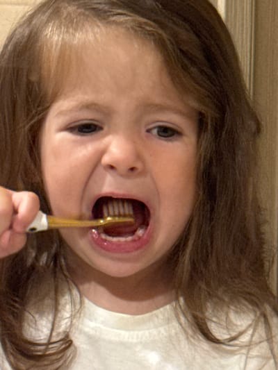 A close-up shot shows a young girl in Beryozovka, Russia, brushing her teeth. She is wearing a white t-shirt and her hair is brown and wavy. She is holding a gold toothbrush in her right hand and it is in her mouth. Her mouth is open and her teeth are visible. The background is blurry, but appears to be a wall.