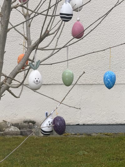 The image shows an Easter decoration, consisting of bare tree branches decorated with colorful Easter eggs. The eggs are pastel-colored with various patterns, including polka dots, stripes, and animal faces. The decoration is located outdoors, with a light-colored wall in the background and green grass visible on the right side of the frame. The location is inferred to be Ampfing, Germany.