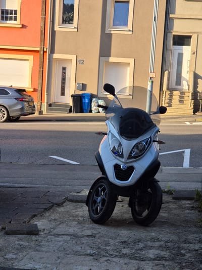 In the city of Sanem, Luxembourg, a white three-wheeled scooter is parked on a sidewalk next to a street. The scooter has a black windshield and mirrors, and a Piaggio logo on the front. The sidewalk is made of broken concrete tiles. Behind the scooter are two trash bins, one black and one blue, and some plastic bags. In the background, there is a two-story building with beige and orange painted walls. The building has several windows and doors, and house numbers "12" and "10" are visible. A silver car is parked in front of the building on the left side of the image. The scene is illuminated by sunlight, casting shadows on the ground and the building.