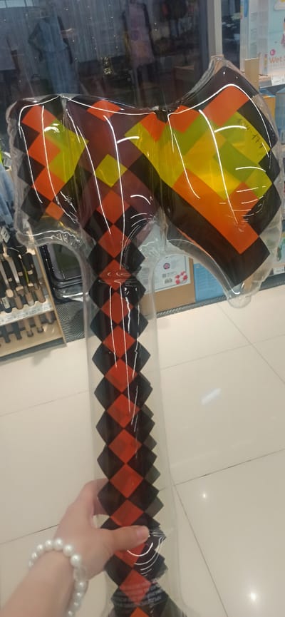 In the city of Asbest, Russia, an individual's hand, adorned with a pearl bracelet, is holding a large, inflatable toy axe. The axe has a diamond pattern with alternating black, orange, and yellow sections on its head, and a black and orange diamond pattern on its handle. The background suggests an indoor retail environment, with shelves stocked with various items, including what appear to be tools or similar merchandise, and a display of clothing or mannequins behind a glass partition. The floor is tiled and appears to be a light color.