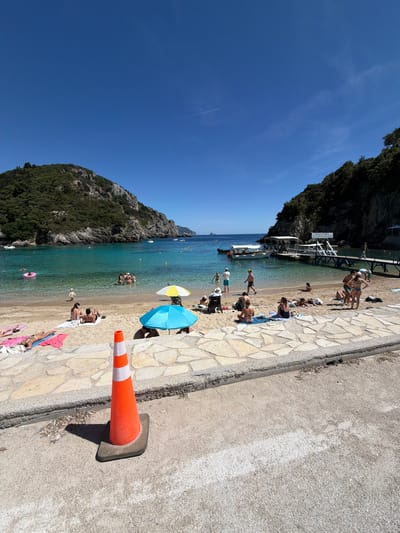 This detailed image depicts a bustling beach scene in Palaiokastritsa, Greece, under a clear, bright blue sky with a faint contrail visible in the upper right. The bay is framed by steep, verdant cliffs with exposed rock faces. The water is a clear, calm turquoise, with yellow buoys marking off a swimming area.

The sandy beach is populated by approximately 25-30 individuals engaged in various recreational activities. On the left side of the beach, several people are sunbathing on colorful towels (pink, white) directly on the sand. A child in a pink inflatable ring is floating in the shallow water nearby. Towards the center, multiple groups of people are standing or sitting. Two beach umbrellas are prominent: one yellow and white, and another solid blue. A wheelchair and a baby carriage are visible on the left side of the beach.

In the water, roughly 10-15 individuals are swimming, standing, or interacting in groups. On the right side of the bay, a wooden pier with a covered section extends into the water. Two white boats are docked at this pier, and several people are seen on and around the structure. A sign with text in Greek is affixed to the pier's shelter.

In the immediate foreground, a textured, light-colored stone-paved walkway transitions to a gray concrete road. An orange traffic cone with two white reflective stripes stands prominently on the concrete road in the lower-center, casting a distinct shadow to its left. The concrete road shows signs of wear, and a faint white line is visible on its surface to the far right. Debris is visible at the junction of the stone paving and the concrete road. The entire scene is brightly illuminated by direct sunlight, creating sharp shadows.