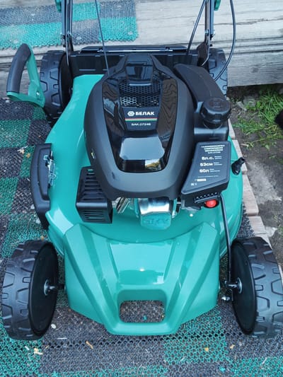 This is an image of a green and black self-propelled gasoline lawnmower, identified by the brand "БЕЛАК" and model number "БАК.07246". The lawnmower is positioned on a patterned mat and what appears to be wooden planks, with some grass and dirt visible in the background. The scene suggests an outdoor setting, likely a yard or garden, with natural lighting indicating it might be daytime.
The lawnmower has a prominent black engine cover with a ventilation grille and a fuel cap. There is a label on the side of the engine that provides specifications: "Мощность 8л.с." (Power 8 hp), "53см" (53 cm cutting width), and "Объём 60л" (60L capacity). A red button, likely a safety or start switch, is visible near the handle. The wheels of the lawnmower are large and black with a tread pattern.
The overall impression is that of a piece of outdoor equipment, ready for use or recently stored. The context of Suvorov, Russia, places it in a specific geographic and potentially seasonal environment. There are no people or animals visible in the image. The weather appears to be fair, with no indications of rain or extreme conditions.