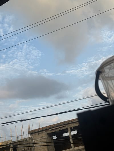 This media file captures a partially constructed building in Amansee, Nigeria, against a sky filled with varied cloud formations. The building appears to be made of concrete blocks with exposed rebar and scaffolding, suggesting ongoing construction. Wooden poles or bamboo sticks jut out from the top of the building. Overhead power lines are prominently visible, crossing the frame diagonally. A part of a translucent, possibly plastic, covering with a dark rim is also in view at the top of the image, obscuring a small portion of the sky. The time of day is difficult to determine precisely, but the lighting suggests it could be late afternoon or early evening, with a mix of sunlight and shadow. The weather appears to be partly cloudy, with patches of clear blue sky interspersed with diffused light and darker cloud masses. There is no visible text in the image. The scene conveys a sense of urban development or renovation in progress.