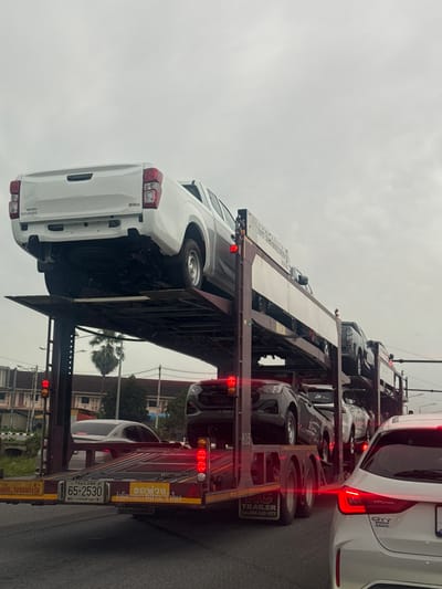 The image depicts a multi-level car carrier truck transporting several vehicles, primarily pickup trucks and sedans, on a cloudy day. The truck is in motion, suggesting it is on a road within a city. The license plate visible on the back of the truck reads "65-2530 THAILAND 91". The environment appears to be urban, with buildings and streetlights visible in the background, consistent with a location in Tambon Si Wichai, Thailand. The truck is large and occupies a significant portion of the frame, with several vehicles loaded onto its upper and lower decks. The sky is overcast, contributing to a subdued lighting condition. The image captures a moment in time of a logistics operation involving vehicle transportation in a city setting.