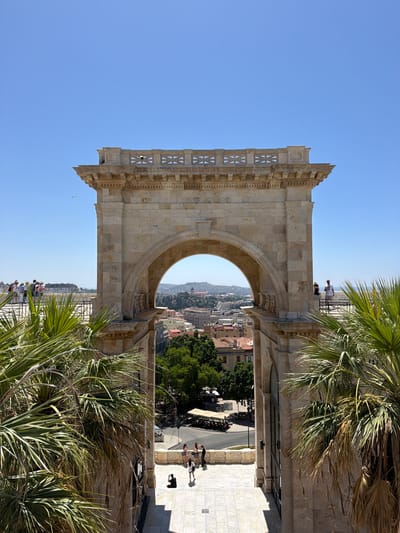 The image shows a sunny day in Cagliari, Italy, with a clear blue sky. The foreground features lush green palm trees on either side, framing an imposing stone archway, likely part of a historical monument. Through the arch, a panoramic view of the city unfolds, revealing terracotta rooftops, buildings with pastel facades, and lush green trees. In the distance, a hilly landscape and hints of the sea can be seen.
Several groups of people are visible, suggesting a tourist area. On a raised terrace to the left, a group of tourists, mostly wearing hats and light clothing, are looking out at the view. On the right side of the arch, two individuals are standing near the railing, also taking in the scenery. Further down, on a paved walkway leading away from the arch, a few more people are scattered, some posing for photos. The overall atmosphere is one of a bright, pleasant day in a popular tourist destination. The architectural style of the arch suggests a historical significance, possibly a triumphal arch or a grand entrance to a significant site.