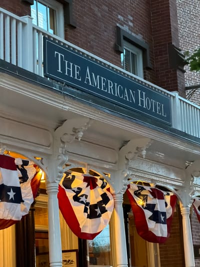 The image shows the facade of The American Hotel, located in the Village of Sag Harbor, United States of America. The hotel has a red brick exterior and white trim. A dark blue sign with white lettering that reads "THE AMERICAN HOTEL" is mounted above a white balcony. Below the balcony, patriotic bunting with red, white, and blue colors, featuring stars, is displayed. The lighting suggests it is either dawn or dusk. No people are visible in the image.