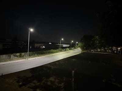 A nighttime street scene in Peshtera, Bulgaria, is depicted, with an asphalt road curving through the frame. Several streetlights illuminate the road, casting pools of light and long shadows. On the left side of the road, a row of buildings with lights on can be seen behind a hedge. The right side of the road features trees and vegetation, with a parked car visible in the distance. The overall atmosphere is dark and quiet, suggesting a residential or suburban area at night.