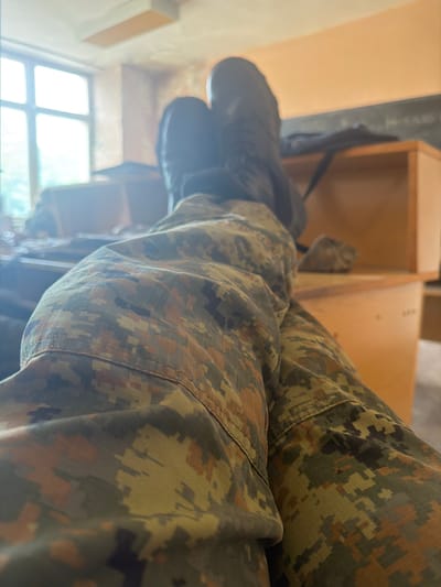 A close-up shot focuses on a person's legs clad in camouflage print trousers. The person is seated. In the background, a window with a light source. The location and city are impossible to be verified.