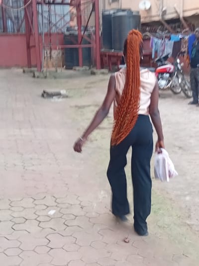 A person with long, orange-brown braided hair wears a light peach top and dark pants. They are walking on a paved surface with a geometric pattern, carrying a white plastic bag. Behind the person is a structure with red metal framing and some black tanks. A motorcycle and people are visible in the background.