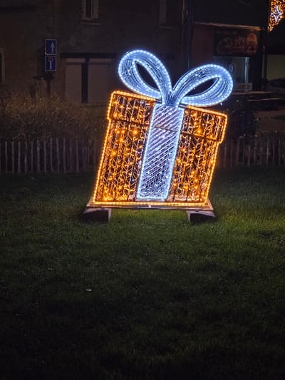 An illuminated decoration depicting a wrapped gift sits on a patch of grass. The gift is boxy and orange with a white stripe. It is topped with a blue bow. Behind the decoration, a building is visible, along with signage. A set of stairs is on the left side of the decoration. The grass is dark and appears to be well-maintained. The scene is likely at night or in low light conditions.
