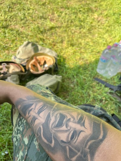The image shows a person's arm with a tattoo of two theater masks, one smiling and one frowning, wearing camouflage clothing, possibly pants. Next to the arm is a helmet, grass, and a plastic water bottle. The specific location in Kostena Reka, Bulgaria cannot be determined from the provided image.