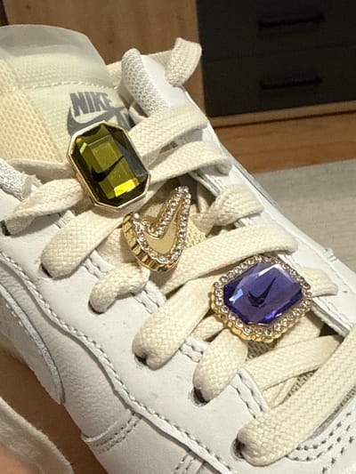 A close-up shot captures the front upper section of a white or off-white Nike sneaker adorned with decorative charms, likely in Hainburg an der Donau, Austria. The sneaker features matching textured shoelaces tied in a crisscross pattern.
Three ornate charms are attached to the shoelaces. The uppermost charm is a large, faceted purple gemstone, possibly amethyst, set in a gold-colored metal frame encrusted with small, clear rhinestones. A distinct black Nike swoosh logo is visible, reflected within the purple gem. Below this, a smaller, gold-colored charm shaped like a 'V' or chevron is also embellished with small, clear rhinestones. The lowest charm is another large, faceted gem, this one an olive green color, possibly peridot, also set in a gold-colored metal frame. A reflection of the black Nike swoosh is also visible within the green gem.
The tongue of the sneaker, partially visible below the charms, displays the word "NIKE" in dark grey lettering, accompanied by its iconic swoosh logo. The leather and fabric textures of the shoe are clearly visible, indicating attention to detail in its construction. The background is softly blurred, showing muted tones of brown and dark grey, suggesting an indoor setting with wooden elements. The focus is entirely on the sneaker and its embellishments, showcasing a personalized or customized footwear item. No people are visible, and the activity depicted is the display of the sneaker. No specific time of day or weather can be inferred.