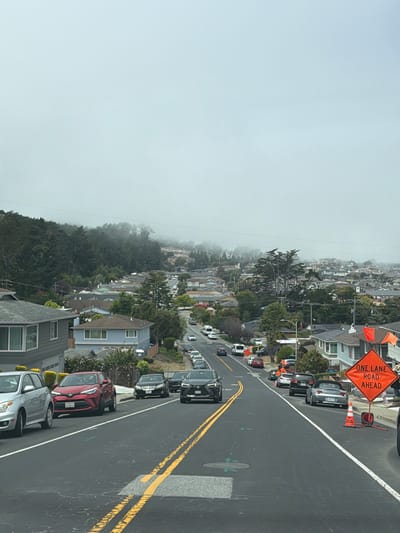 The image depicts a residential street in San Bruno, California, USA, with a hilly landscape and dense fog. The street is lined with parked cars and houses, and a traffic cone and "ONE LANE ROAD AHEAD" sign are visible on the right side of the road. Several cars are driving up the hill, and some pedestrians are visible in the distance. The overall atmosphere is overcast and somewhat hazy due to the fog.