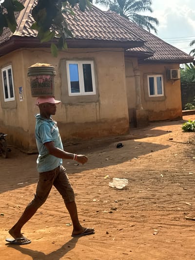 A male subject is depicted walking left-to-right on a reddish-brown dirt path in Aguata, Nigeria, under direct sunlight. He wears a light blue short-sleeved collared shirt, dark brown knee-length shorts, light brown flip-flops, and a pink baseball cap. A dirty white plastic bucket, marked with pink and black text including "GOLDEN HORSE," containing a tan granular material, is balanced on his head over the cap. Red and white bracelets are visible on his right wrist. In the background stands a single-story, light brown building with a brown tiled roof. It features multiple white-framed windows with blue reflective glass, and an exterior white air conditioning unit is mounted on the wall to the right of one window. The ground exhibits tire tracks and scattered refuse, including a crumpled white plastic bag. Green foliage is visible in the upper left and behind the structure. Shadows are cast prominently to the left.