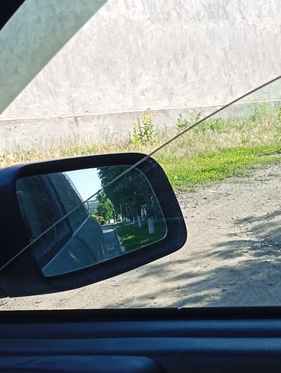 The image shows a side-view mirror of a car, reflecting a rural road lined with trees and houses. The mirror itself is cracked, with visible lines disrupting the reflection. Through the car window, a textured concrete wall is visible, casting a shadow from an unseen object. The scene appears to be set in a rural location, possibly a village, with a dirt road and vegetation. The time of day seems to be daytime, with bright sunlight creating shadows. The sky is clear and blue in the reflection. There is no visible text in the image. The overall impression is that of a casual snapshot taken from inside a moving vehicle, capturing a glimpse of the surrounding environment.