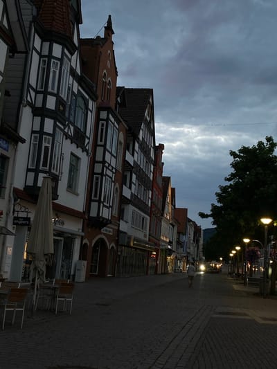The image depicts a street scene in Rinteln, Germany. The architecture of the buildings lining the street is predominantly traditional half-timbered style, with white stucco and dark timber beams. The buildings have steeply pitched roofs, many with multiple gables and dormer windows. The street itself is paved with cobblestones.
In the foreground on the left, there are outdoor tables and chairs belonging to a cafe or restaurant, covered by a large, light-colored umbrella. Signs for "engbers" and "NEUERÖFFNUNG" are visible on the storefronts.
Further down the street, a single person is walking away from the camera. A vehicle with bright headlights is approaching from the distance, casting a strong glow. Streetlights with hanging flower baskets illuminate the right side of the street, and trees line this side as well. The sky is overcast with dark, cloudy conditions, suggesting it is late afternoon or early evening. The overall impression is of a quiet, historic European town street.