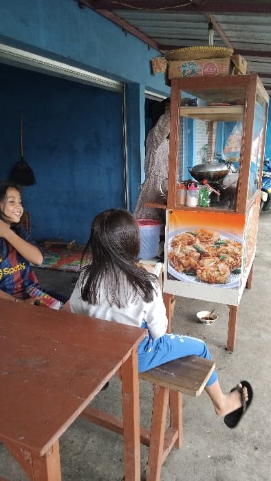 The image, taken in Panjalu, Indonesia, captures a bright daytime scene outside what appears to be a street food stall or small shop. Two young individuals are present, positioned near a wooden food cart.
In the foreground, a girl with long dark hair, wearing a white long-sleeved top and blue pants, is seated on a wooden stool with her back mostly towards the camera. Her right foot, clad in a black sandal, is visible resting on another part of the wooden structure. To her left and slightly lower, another girl is visible, smiling warmly and looking upwards. She wears a blue and red patterned shirt with a "Spotify" logo visible on her sleeve. Her arm is around the lower back of the first girl, suggesting a friendly interaction.
Central to the image is a wooden food cart with a glass display case, likely used for selling food. The cart features a prominent advertisement on its side displaying an image of several golden-brown fried fritters, commonly known as Bakwan in Indonesia. Inside the cart, a dark wok or pan and various containers are visible, indicating food preparation or serving. To the far left of the cart, a woven bamboo tray and a cardboard box, possibly containing "Mi Sedap" instant noodles, are placed on a blue surface.
The setting includes a concrete ground and a blue wall or structure behind the people and the food cart. A small white bowl, possibly used for dipping sauce or drinks, is seen on the ground near the first girl's feet. The overall impression is one of a casual, lively local food vendor scene. The bright lighting suggests it is daytime.