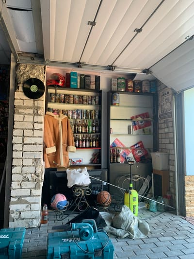 The image shows a cluttered garage in Afanasovo, Russia, with a partially open garage door revealing a glimpse of the exterior. The scene is filled with various items, including a red motorcycle helmet, shelves stocked with bottles of alcohol and cans, a tan sheepskin coat hanging on a rack, a basketball, a pink and blue ball, a glass bottle with an amber liquid, a bright yellow garden sprayer, several glass jars, and two teal power tool cases labeled "Makita" in the foreground. A black vinyl record is mounted on a white brick wall to the left. The floor is paved with gray bricks.