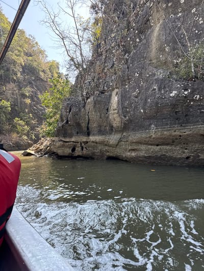 The image shows a close-up view from a boat moving along a rocky shoreline. The rock face is layered and textured, with some vegetation clinging to it. Dark green water is visible, churned by the boat's movement creating white foam. A portion of the boat, including a red life jacket, is visible in the foreground. The photo is taken on a sunny day.