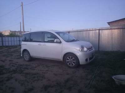 In the city of Kenjekol, Kazakhstan, a white van is parked on a dirt and grass area. The van is facing left and is positioned in the center of the frame. Behind the van is a corrugated metal fence, and to the left of the van, a utility pole with wires is visible. In the background, a brick building with a dark roof is partially obscured by the fence. A green hose lies on the ground in front of the van, and a light-colored bucket is visible to the right of the hose. The sky is a clear blue, indicating it is daytime.