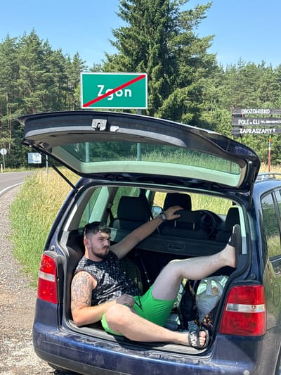 A man is sitting in the open trunk of a dark blue station wagon. He is wearing a black and white patterned t-shirt and bright green shorts. He has a tattoo on his left arm. His legs are extended out of the trunk. The car is parked on the side of a road with a grassy verge. In the background, there are trees and a blue sky. A green sign with a red diagonal line through the word "Zgon" is visible on the left side of the image. On the right side, a wooden sign with text appears to be for a campsite. The man is looking to his right.