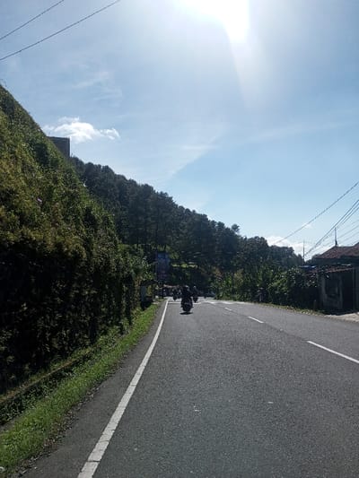 The image displays a two-lane asphalt road under a bright, sunny sky with minimal cloud cover in Ka Thaung (Upper), Myanmar. The sun is prominently featured in the upper right quadrant, emitting a strong glare. To the left of the road, a steep hillside rises, densely covered in green vegetation and trees. A dark, rectangular structure is partially visible near the top of this slope. The road itself has a solid white line marking the left shoulder and a dashed white line indicating lane separation further into the distance. Several dark-colored motorcycles are observed traveling away from the viewer along the road, with one in the immediate foreground carrying a single rider. In the mid-ground, a large billboard displaying an advertisement is suspended over the road amidst abundant foliage. To the far right of the road, a weathered building with a red-tiled roof and light-colored walls is situated. Power lines traverse the upper portion of the frame and are also visible above the building on the right. A narrow grassy verge and a drainage ditch run alongside the left edge of the road.