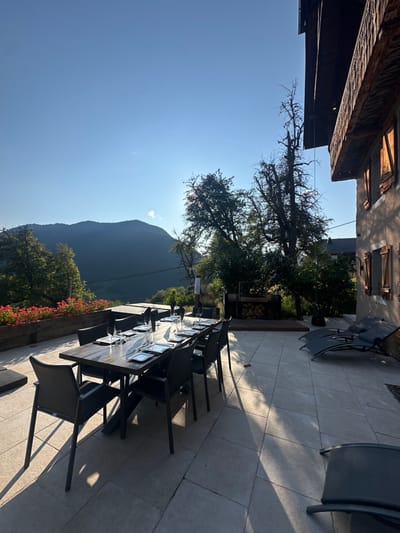 The image depicts an outdoor patio scene in Saint-Jean-d'Aulps, France, at what appears to be late afternoon or early evening, indicated by long shadows cast across the light-toned square patio tiles.
In the foreground and midground, a long, rectangular dark wood dining table is centrally positioned, set with eight dark chairs. Each place setting includes a dark square plate, a folded white napkin, a wine glass, and flatware. Beyond the table, to the left, a wooden planter box filled with vibrant red flowers runs along the patio edge.
To the right of the dining table, two dark recliners or loungers are positioned on the patio. Further right, a multi-story building with light-colored walls and dark wooden window shutters, some appearing open, occupies the extreme right edge. A wooden balcony with intricate detailing extends from the upper floor of this building.
In the background, a series of dark, forested mountains are visible under a clear, bright blue sky, with the sun low on the horizon, partially obscured by trees to the right. Directly behind the dining table and adjacent to the building, a large, tall, leafless tree stands prominently. Below this tree, on a raised wooden platform, an outdoor cooking or grilling station is visible, accompanied by a stack of firewood. A faint, blurry human figure is discernable near this area. The overall environment suggests a tranquil, mountainous setting.