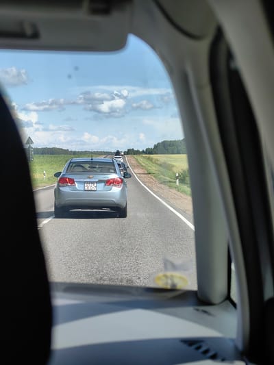 The image shows a view from inside a vehicle, looking out through the windshield and side window onto a two-lane road. The road curves slightly to the right, with a field of green crops and trees lining the left side, and a grassy verge with a white post on the right. A line of cars is visible on the road ahead, traveling in the same direction. The lead car, a light blue Chevrolet Cruze, is the most prominent subject. Its license plate, which appears to be from Belarus, reads "0179 KO-7". The sky is blue with scattered white clouds. The interior of the vehicle, including a portion of the sun visor, the edge of the windshield frame, and part of the driver's side interior panel, are visible in the foreground. The scene appears to be a rural setting in the vicinity of Хаценчыцкі сельскі Савет, Belarus, likely on a sunny day.