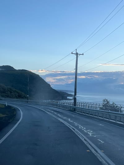 The image displays a coastal road at dawn or dusk, identified as the Sea Cliff Bridge near Stanwell Park, Australia. In the immediate foreground, a dark asphalt road curves slightly to the left, marked with a solid white line on the left edge and double solid white lines centrally, interspersed with yellow reflective road studs. Faint, weathered white markings are visible on the road surface to the right of the double lines. To the right of the road, a silver metal barrier with multiple horizontal rails separates the road from the ocean.

A tall, weathered wooden utility pole with multiple cross arms and numerous power lines extending horizontally across the frame stands immediately adjacent to the barrier on the right. Another, smaller utility pole is visible further along the road on the left, partially obscured by the landscape.

Beyond the barrier, the calm, grey-blue ocean extends to the horizon, with gentle waves visible near the shore. The coastline features dark, rugged cliffs meeting the water. In the distance, a distinct rectangular aperture or tunnel entrance is visible within a cliff face directly over the ocean.

To the left, a steep, densely vegetated green hillside rises sharply, exhibiting some wisps of mist near its crest. The sky above is a clear, light blue, transitioning to a paler shade near the horizon, with a horizontal band of layered clouds displaying soft pink and orange hues along their undersides. The overall lighting is soft and diffuse. No individuals or active subjects are present in the scene.