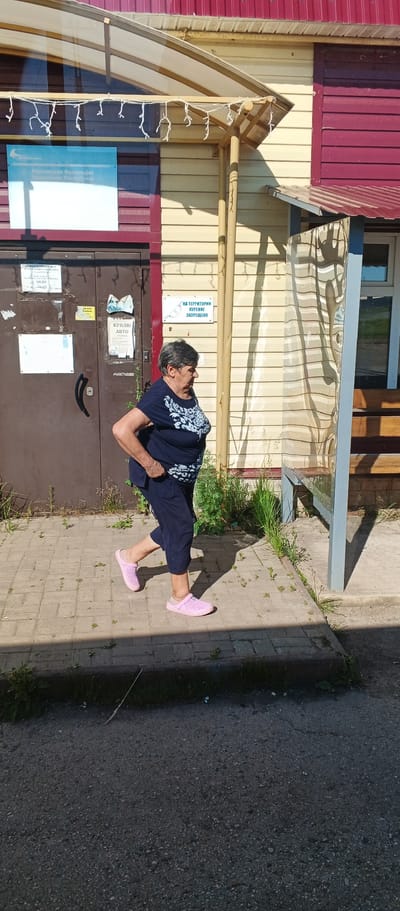 An older woman with short gray hair, wearing a dark blue shirt with a white pattern and dark blue cropped pants, walks on a paved sidewalk. She is wearing pink slip-on shoes. She is walking from left to right in the frame, with her body angled towards the right. In the background, there is a building with yellow and red siding. There are several signs on the building, including one with Russian text that reads "НА ТЕРРИТОРИИ КУРЕНИЕ ЗАПРЕЩЕНО" which translates to "SMOKING IS PROHIBITED ON THE TERRITORY." There is also a glass-enclosed bus stop or waiting area to the right of the building. The lighting suggests it is daytime, with shadows cast on the ground. The overall scene appears to be an outdoor area adjacent to a building in a residential or commercial district. This scene is located in the village of Kiyasovo, Russia.
