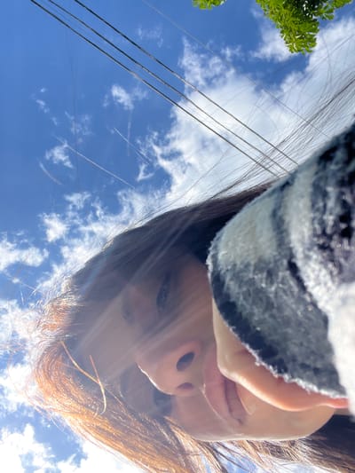 The image is an upward-facing perspective shot, featuring a person's face in the upper right quadrant, with brown and possibly auburn hair catching the sunlight. The individual is wearing a black and white striped garment. The background consists of a bright blue sky with scattered white clouds. In the upper left, there are some green leaves. The presence of power lines running across the sky suggests an urban or suburban setting. Without additional elements or context, determining the specific location, particularly in Blizhniy Khutor, Moldova, is impossible.