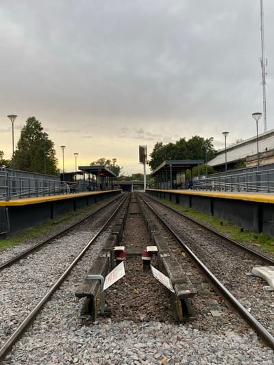 The image displays a train station in Sáenz Peña, Argentina, featuring parallel railway tracks and platforms under an overcast sky. The photograph is taken from near track level, looking down the length of the tracks as they recede into the distance.
On the right side of the image, two sets of railway tracks are clearly visible, laid on a bed of grey gravel ballast and wooden sleepers. A third rail or similar component runs parallel to the main rails. Attached to one of the wooden sleepers is a small white sign with red lettering, partially obscured, that reads "PELIGRO" (Spanish for "DANGER").
To the left of the tracks, a long platform stretches into the background, distinguished by a bright yellow safety strip along its edge and a metal railing. Further along this platform, there is a covered waiting area or shelter where indistinct figures of people can be seen. Underneath the shelter, a red digital display is visible, likely indicating train information. Several lamp posts with uniquely shaped lights are spaced along the platform. Trees and other foliage grow alongside the platform and in the background. In the far distance, additional station structures and a tall communication or power tower are visible against the sky.
The sky is predominantly overcast, but a warm, yellowish glow emanates from the horizon in the distance, suggesting either early morning or late afternoon light. The overall lighting is soft and diffused. The scene depicts a quiet, functional train station during a transitional period of the day.
