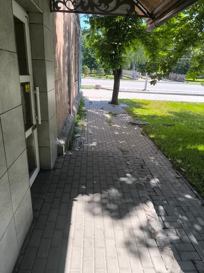 The image shows a sidewalk next to a building and a street in the city of Bohodukhiv, Ukraine. On the left is the side of a building with a door and a yellow sign that says "OFFERT". The sidewalk is paved with rectangular gray bricks. To the right of the sidewalk is a strip of green grass and then a street. There is a tree on the sidewalk, and its shadow is cast across the bricks. In the distance, there are more trees and some buildings. The weather appears to be sunny, as indicated by the strong shadows. There are no people visible in the image.