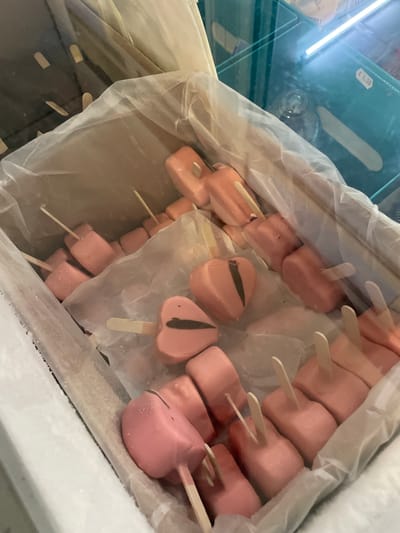 The image shows a close-up, high-angle view of a chest freezer containing numerous pink, heart-shaped and rectangular popsicles on sticks. The popsicles are arranged in a chaotic manner, some partially covered by white paper or plastic. The freezer is lined with white plastic. In the background, a glass display case is visible, with some items inside and a price tag of "€ 1.50." The scene appears to be an interior view of a commercial establishment, likely a shop or kiosk, possibly in Skala Avdiron, Greece, given the presence of Euros. There are no people visible in the image.