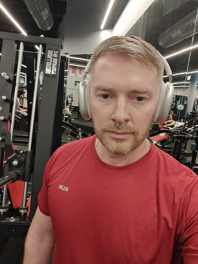 An adult male is depicted in a close-up selfie taken inside a gymnasium located in Nahariya, Israel. The subject, a fair-skinned individual with short light brown hair, a ginger-colored beard, and light eyes, is wearing a red short-sleeved athletic shirt with the word "RUN" printed in white on the left chest, appearing reversed due to reflection. He is also wearing silver over-ear headphones and a silver chain around his neck. The background reveals gym equipment, with a black cable machine partially visible to the left. A large mirror behind the subject reflects more exercise machinery, including a leg press with an individual seated on it, other gym patrons, and red lettering, possibly "LUX," on the mirrored wall. The ceiling features linear overhead lighting and air conditioning vents.