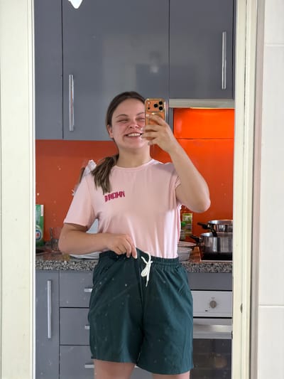 A young woman is taking a selfie in a bathroom mirror. She is wearing a pink t-shirt and green shorts. The bathroom has gray cabinets and an orange backsplash. A section of what appears to be a kitchen is visible in the background with stainless steel appliances. The location is Arroyo de la Miel-Benalmádena Costa, Spain.
