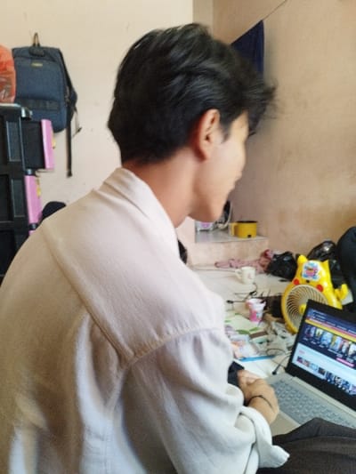 An indoor scene in Muka, Indonesia, depicts a young individual, likely male, from a slightly elevated rear-right perspective. The individual, possessing dark hair, wears a light-colored, long-sleeved collared shirt and is seated, facing towards the right side of the frame, engaged with an open silver/gray laptop. The laptop screen displays a web page featuring multiple small video thumbnails. To the right of the laptop is a small yellow desk fan and, behind it, a partially visible yellow inflatable object. A white mug and a pink-patterned cup sit on the surface near the laptop, surrounded by tangled cables and wires. In the background, to the left, a dark blue backpack hangs from a hook above a dark-colored, multi-tiered storage unit with pink accents. Further back and to the right, a small yellow pot rests on a raised, tiled platform. A dark blue fabric hangs on the light brown/beige wall in the upper right quadrant. The tiled floor in the mid-ground shows scattered loose items. The overall environment suggests a cluttered domestic setting.