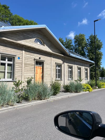 In Tartu, Estonia, a long, light-colored wooden building with a blue metal roof stands under a clear blue sky with a few white clouds. The building features multiple white-framed windows and a single door, which is painted bright orange with what appears to be orange tape or paint marks applied in a grid pattern. The facade is constructed of horizontal wooden planks, and decorative molding is visible above the windows. A small, semi-circular window is situated in the gable. In front of the building, a well-maintained garden bed contains tufts of tall, feathery grey-green grass and bright yellow-green leafy plants. The garden is bordered by a concrete curb and a gravel path. A modern black lamppost stands to the right of the building, and trees with lush green foliage are visible in the background. The foreground shows an asphalt road and the side mirror of a vehicle, reflecting the road and the surrounding environment.