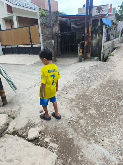 A young boy stands on a cracked and uneven concrete surface in what appears to be a residential alleyway. He is wearing a yellow jersey with "RONALDO" and the number "7" on the back, along with blue shorts and black sandals. The background features houses with a mix of brick and painted walls, some with gated entrances and overgrown vegetation. Power lines and a cloudy sky are visible above.