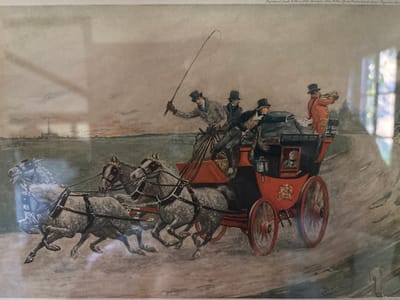The image is a photograph of a framed historical print, depicting a four-horse mail coach in rapid motion across an open landscape. Reflections on the print's surface indicate it is displayed under glass.

The central subject is a vibrant red and black four-wheeled coach, angled from the lower-left foreground, moving towards the right background. It is drawn by four light-colored (grey or white) horses, each equipped with dark harnesses, shown mid-gallop, conveying speed and dynamism.

Five individuals are prominently visible on the coach. The driver, seated on the elevated front bench, wears a dark top hat and light jacket, holding reins and a long whip raised in his right hand. Two additional male passengers in dark top hats and coats are seated next to him. At the rear of the coach's upper deck, a man in a bright orange-red jacket and dark top hat holds a long brass horn to his mouth. Through a small, dark-framed window on the coach's side, a partial view of a passenger is discernible. The side of the coach bears a gold crest and the word "LONDON" in white lettering on a dark panel.

The environment features a dirt or unpaved road, showing tire tracks and implied movement. The background to the left comprises flat green fields extending to a distant horizon, where a cluster of buildings, including a tall smokestack or tower, is visible. The right side of the road is bordered by sparse foliage and a faint wooden fence line. The sky is overcast, displaying a gradient of grey and pale brown tones.

Text in the upper-right of the print identifies the publisher as "Raphael Tuck & Sons Ltd. London, New York. Publishers to Her Majesty the Queen." A partial artist's signature is present in the lower right. The depicted scene, a historical four-horse mail coach, is identifiable as being set in Great Britain due to the inscription "LONDON" on the coach and the British publisher's details. This particular print, as photographed, is located in the city of Monterey, United States of America.