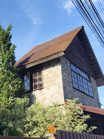 The image depicts a two-story building located in Быково, Russia, viewed from a low angle on a clear day with a bright blue sky and minimal wispy clouds. The building's walls are constructed from light-toned, possibly concrete or cinder blocks with visible mortar lines, showing signs of weathering. A steep gable roof, covered with corrugated reddish-brown metal sheets, exhibits extensive rust and oxidation. The gable ends are clad with dark horizontal wooden planks.

On the building's left side, two windows with dark wooden frames and multiple panes are present; the leftmost window is open outwards. On the right side, a larger, multi-paned window with a dark wooden frame is visible. A dark brown rain gutter or awning extends below the large window.

In the left foreground, a tall evergreen tree with dense green foliage partially obscures the building. In the lower midground and foreground, variegated green and white bushes are prominent, partially concealing a dark brown corrugated metal fence. An orange, dome-shaped light fixture is mounted on a post in the bottom right foreground. Several dark, parallel utility lines or cables diagonally traverse the upper right quadrant of the sky. Bright sunlight illuminates the scene, casting shadows consistent with a clear day.