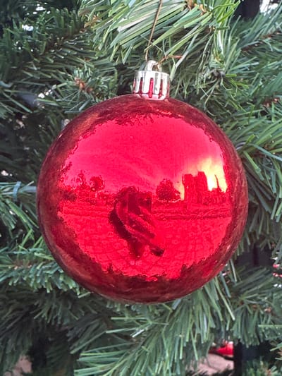 The image presents a close-up view of a single, bright red, spherical Christmas ornament hanging on what appears to be an artificial Christmas tree. The ornament's surface is highly reflective, capturing distorted images of its immediate surroundings.

Within the prominent reflections on the glossy red surface, several details are discernible. A central, elongated, somewhat shadowy form resembles a hand or arm, possibly belonging to the person holding a camera or phone to capture the image. Around this, there are bright, undefined shapes and rectangular forms that could be windows, light sources, or other elements of an indoor environment. A strong, bright yellowish reflection is visible towards the upper left side of the ornament. The ornament itself has a slightly textured or possibly glitter-infused finish, which subtly diffuses the reflections.

The ornament is secured by a silver cap at its top, from which it hangs among the dense, deep green needles of the artificial tree, forming the blurred background. There is no explicit activity shown, but the reflections implicitly indicate the presence of a photographer. The scene suggests an indoor setting, likely well-lit. No text is visible in the image. The location context for this photograph is Agios Dometios, Cyprus.