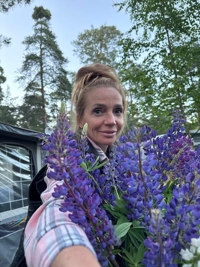 A woman with blonde hair pulled into a bun is holding a bouquet of purple lupine flowers in the foreground. She is wearing a pink and white plaid shirt and a dark jacket. Her left arm is visible, holding the flowers. In the background, there is a tent and trees. The sky is visible above the trees. The image appears to be taken outdoors, possibly in a campsite or a park. The location is specified as Ozerki (Krasnodolinskaya), Russia.