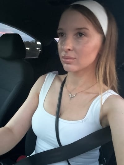 A young woman with blonde hair and a white headband is sitting in the driver's seat of a car, looking to the left. She is wearing a white tank top and a delicate necklace with a pendant. A black seatbelt is across her chest. The car's interior is dark, with the passenger seat visible behind her. Outside the car window, a twilight sky with hints of pink and blue can be seen, along with distant lights and trees. The overall impression is a snapshot of a woman on a drive during dusk in Petukhovo, Russia.