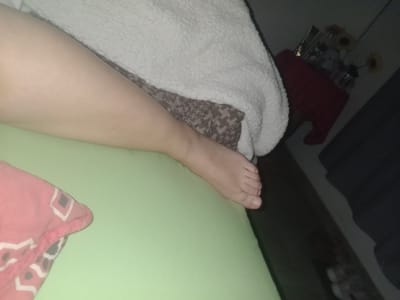 A woman's leg lies on a bed with green sheets and a patterned blanket. The foot is visible in the upper left portion of the image. To the left, there is a shelf with a dark finish and a red item partially visible. Based on the description, the location is likely in Villa Ballester Noroeste, Argentina.