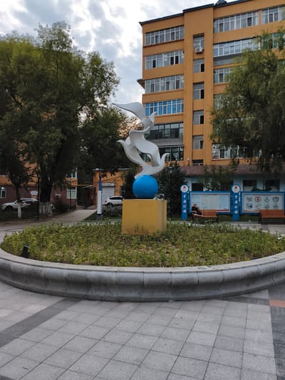 In Jixi, China, a white sculpture of two stylized birds is positioned in the foreground, appearing to take flight from a bright blue sphere. The sculpture rests on a yellow rectangular pedestal, which is surrounded by a circular planter filled with low-lying green foliage. A grey, curved concrete border encircles the planter. Beyond the sculpture, a tree with dense green leaves partially obscures a building. The building is a tall, orange structure with numerous windows, some of which have blue tinting. To the right of the building, a section of a pale blue structure with display boards is visible. A man in dark clothing sits on a wooden bench in front of these display boards. Two white cars are parked on a paved area behind the planter and to the left of the building. The ground in the foreground is paved with grey square tiles. The sky above is overcast with clouds.