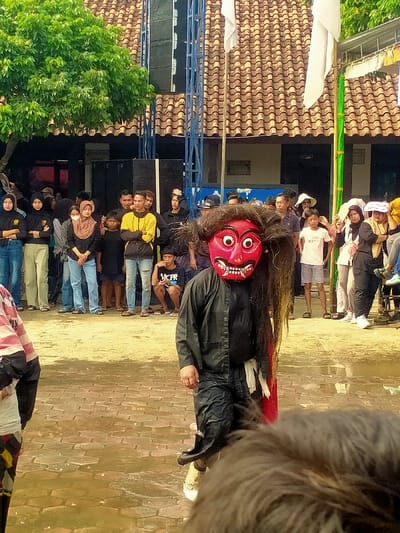 The image depicts an outdoor event in Tamansari, Indonesia. A central figure in the mid-foreground wears a large, ornate red mask featuring prominent white eyes, an open mouth with visible teeth, and long, dark, shaggy hair. This individual is dressed in a black long-sleeved button-down shirt, dark trousers, and light-colored athletic shoes, with a red fabric section and a white fabric element visible on their left side. The immediate ground is a wet, light-brown paver surface. Behind the masked figure, approximately twenty-five onlookers are gathered, primarily standing. This crowd comprises adults and children, with several women wearing hijabs and some individuals in black shirts with yellow emblems. In the background, a building with a multi-layered reddish-brown tiled roof and a dark, recessed ground floor extends across the scene. To the left, a large green tree is present. Two blue metal truss structures, one fitted with speaker equipment, stand against the building. Two white flags are affixed to poles on the right side of the background. The upper portion of a person's dark, shaggy head is visible in the lower right foreground.