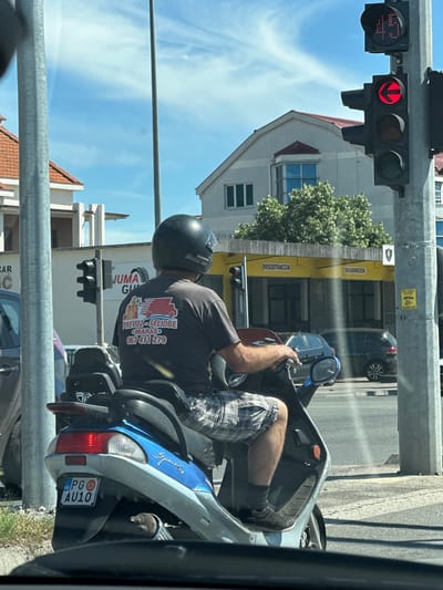 In Pelev Brijeg, Montenegro, a man wearing a black helmet rides a blue and silver scooter. He is wearing a dark gray t-shirt with a red and white logo and text on the back, and plaid shorts. The scooter has a license plate that reads "PG AU10". He is stopped at a traffic light that shows a red left arrow and a digital display showing "45". In the background, there are buildings, trees, and other vehicles. The traffic light is mounted on a metal pole. The weather is clear and sunny.